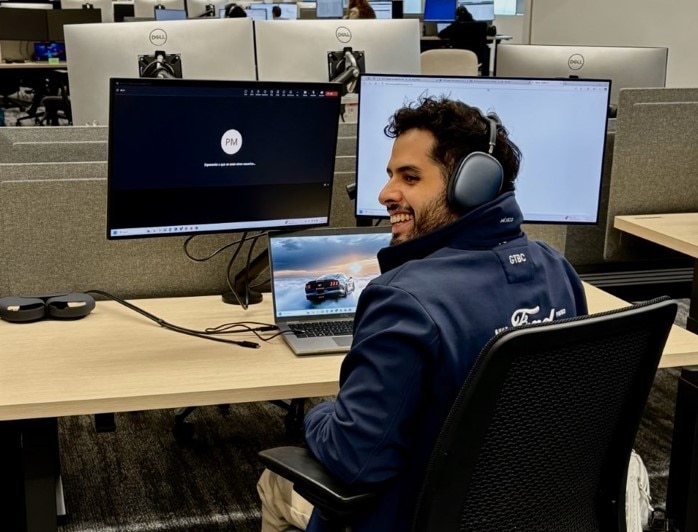 A man wearing headphones sits at a desk, focused on two computers in front of him.