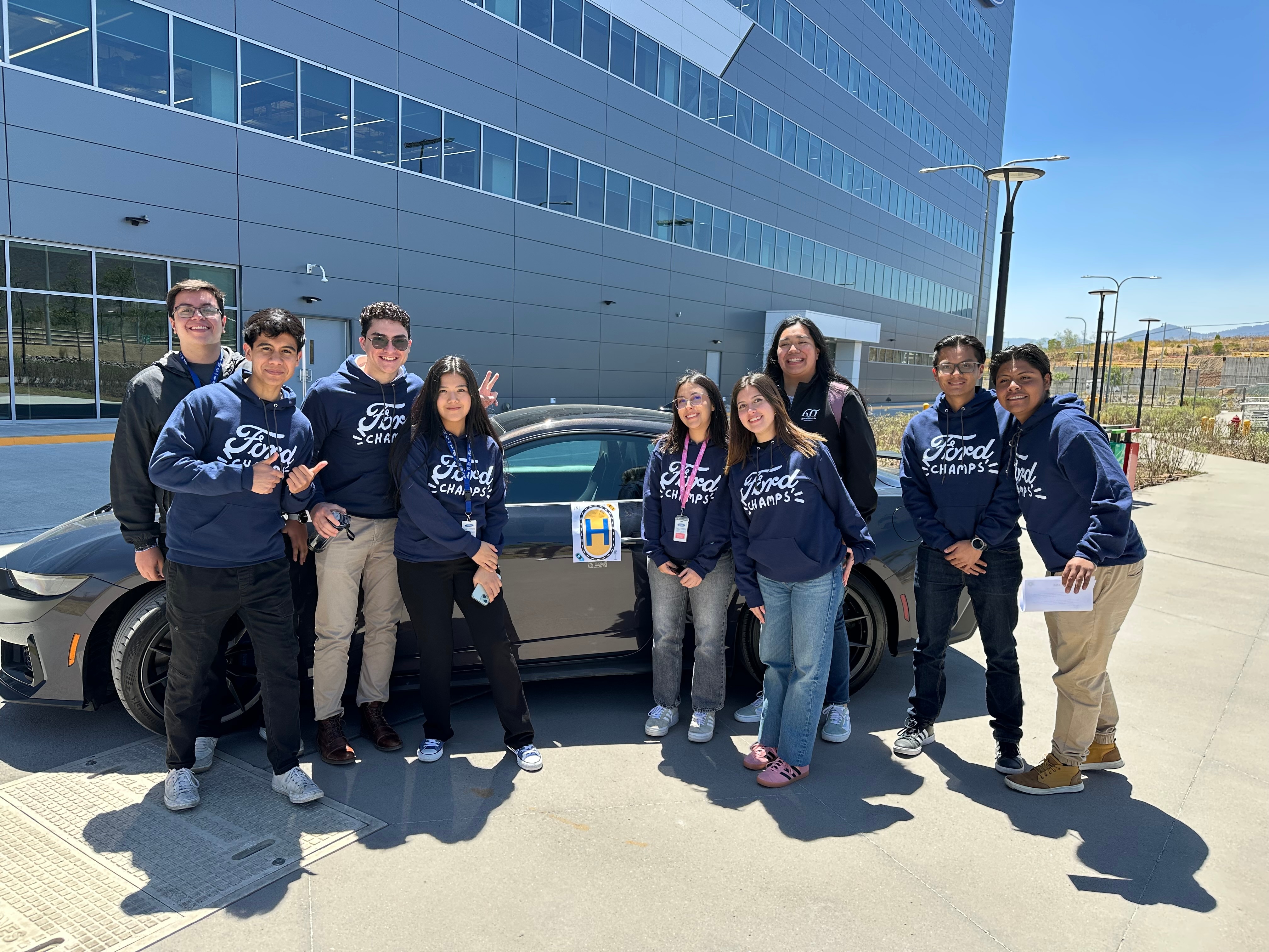 A group of students smiling and posing together in front of a parked car on a sunny day.