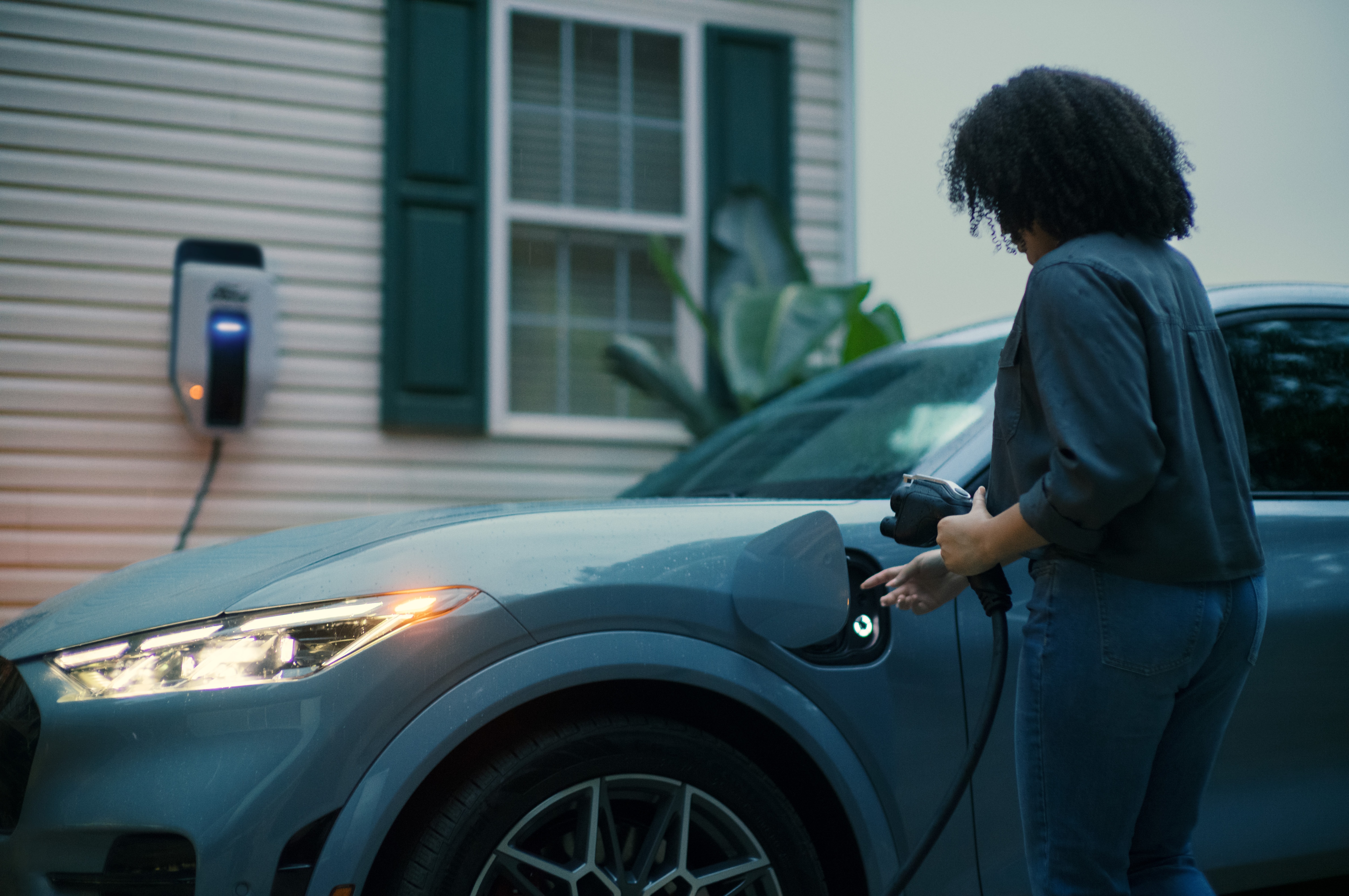 A woman is charging her electric car at a home charging station in a residential driveway.
