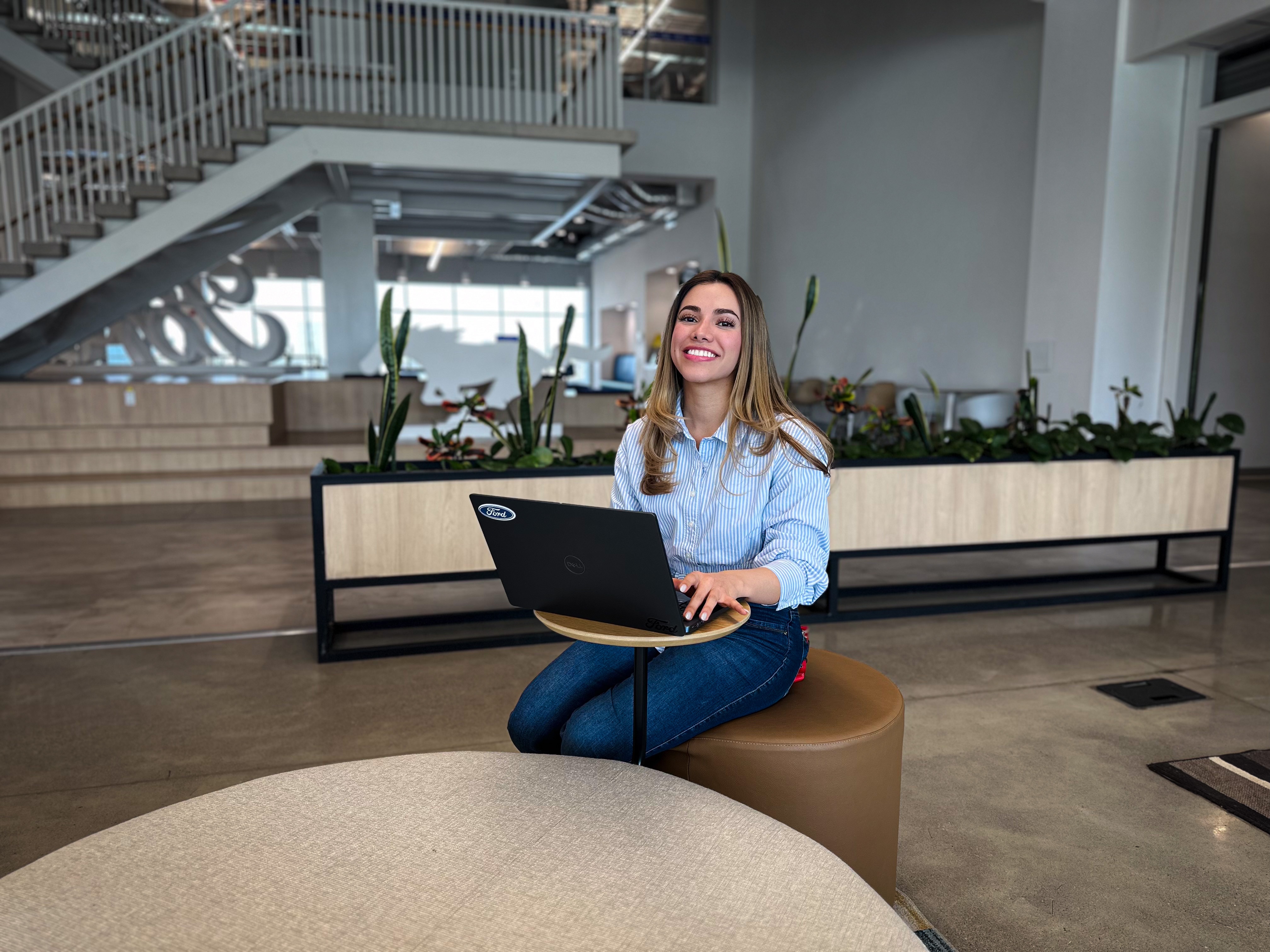A woman sitting on a stool, focused on her laptop, with a neutral background.
