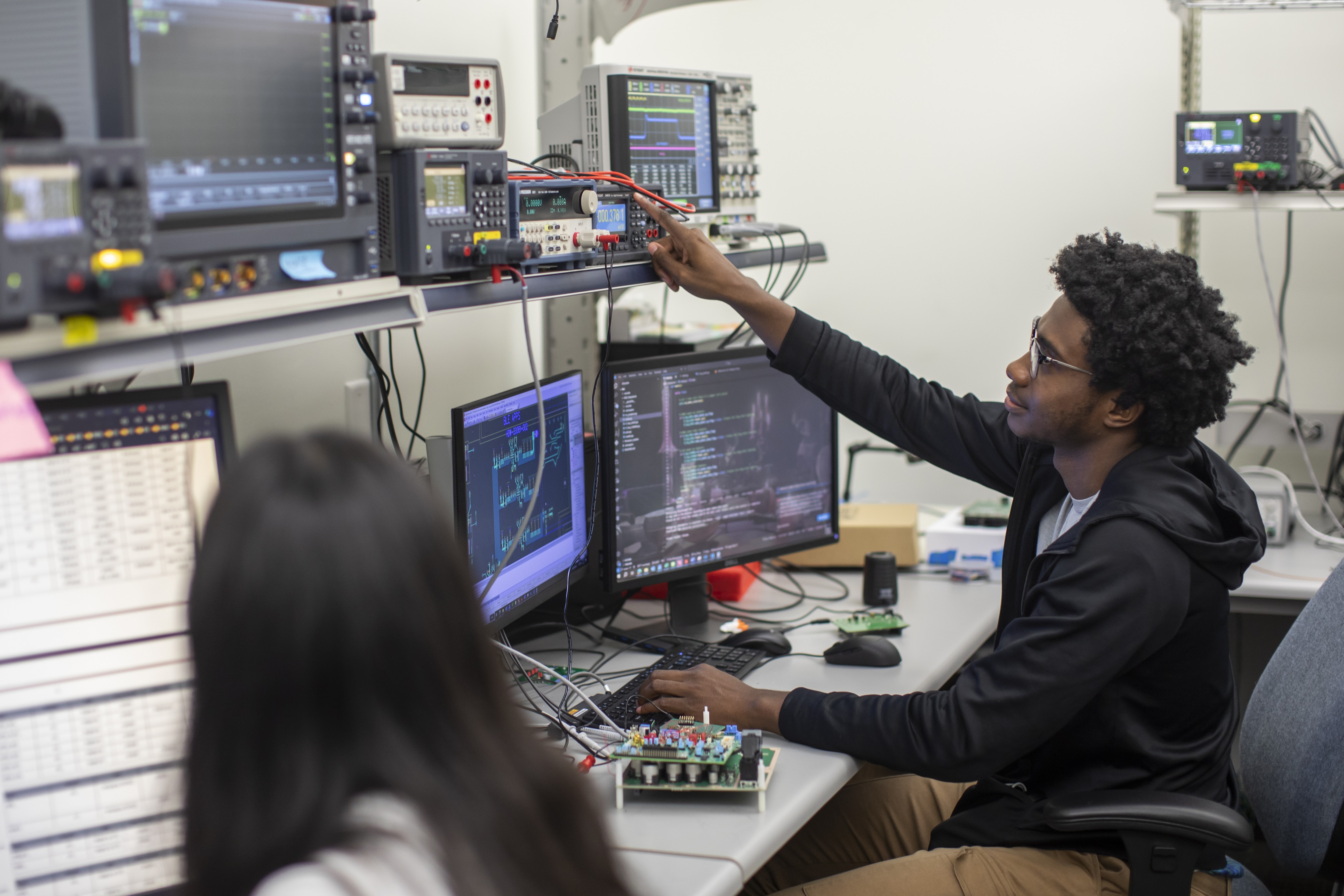 Two people work in a lab with computers and electronic equipment. One person adjusts a device on a shelf while the other is blurred in the foreground.