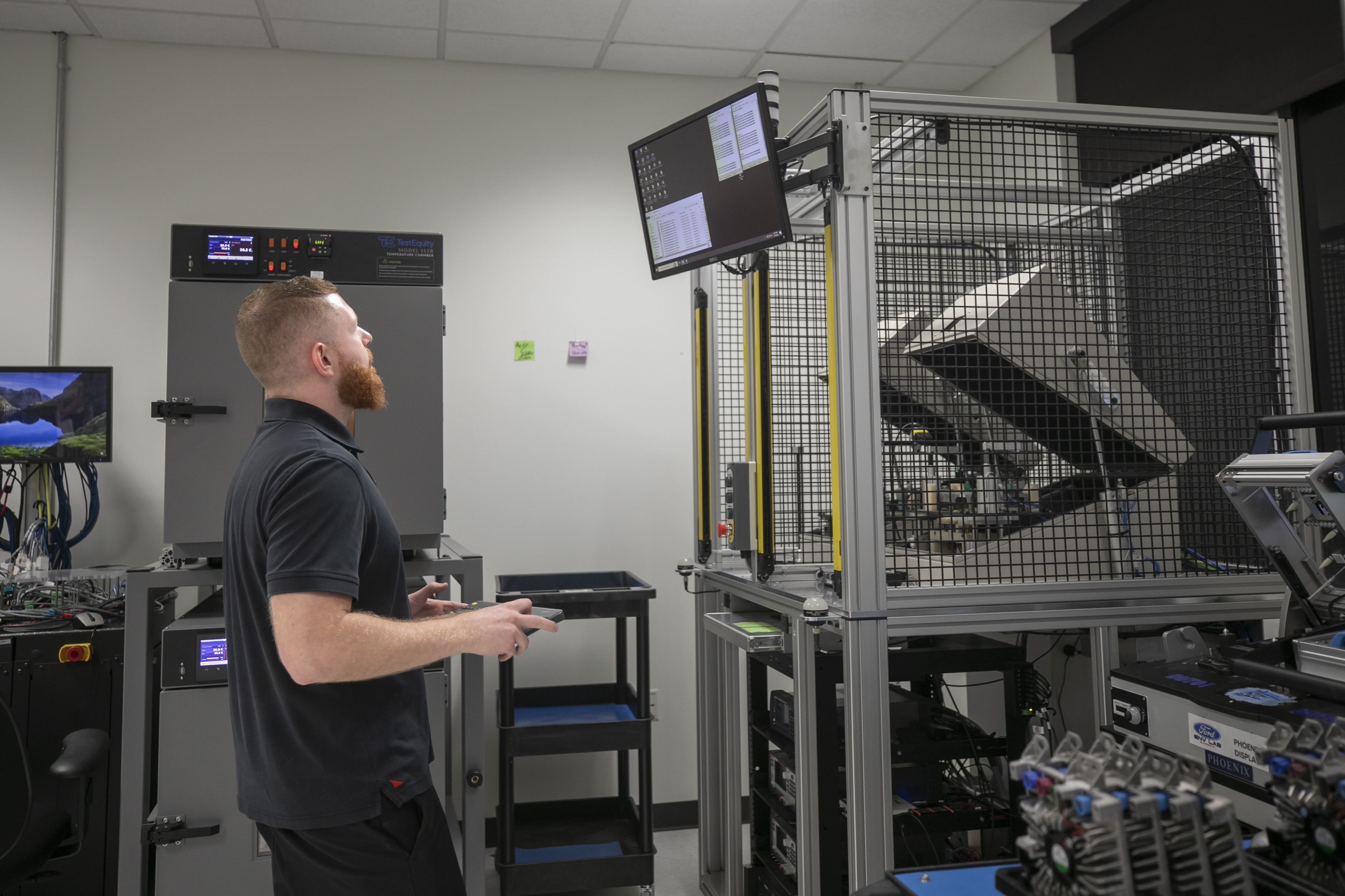A person stands in a tech lab, observing data on a monitor. The room contains electronic equipment and metal frameworks.